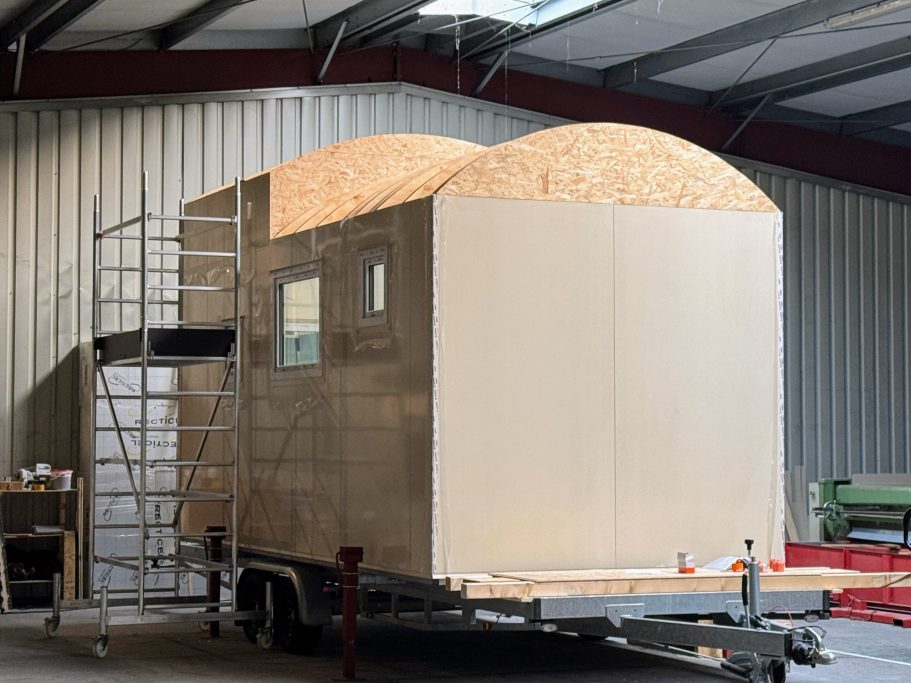 Teilfertiges Tiny House in der Produktionshalle auf Trailer. Ein Gerüst steht daneben.