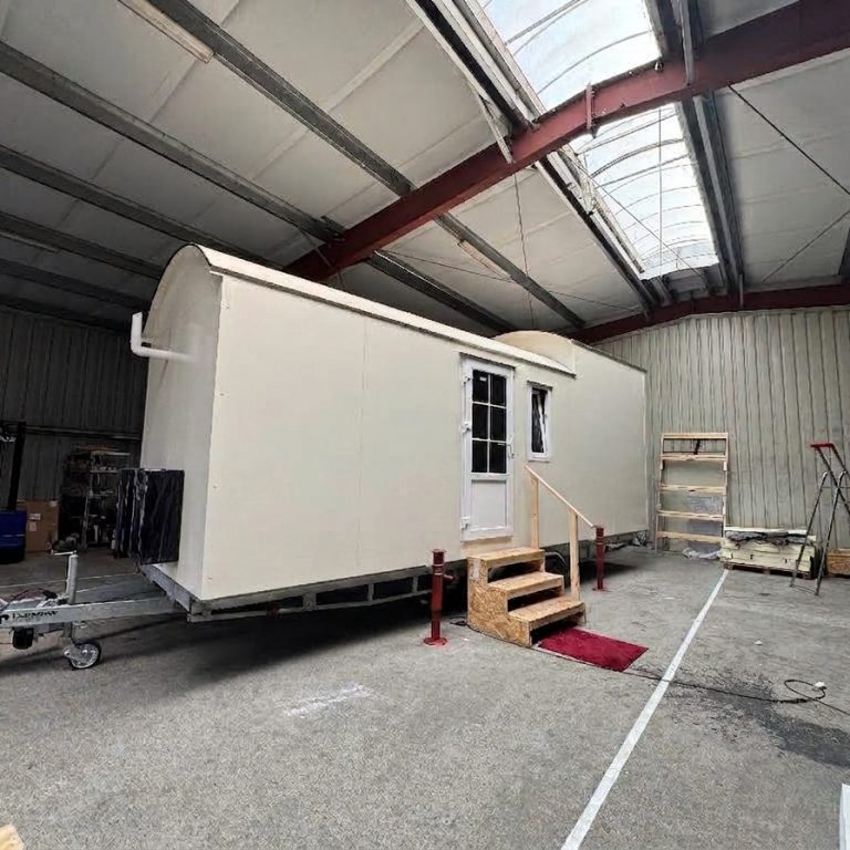 Einfarbiges Tiny House mit schwarzen Boxen und weißem Schornstein in Produktionshalle auf Trailer. Treppe mit rotem Teppich davor.