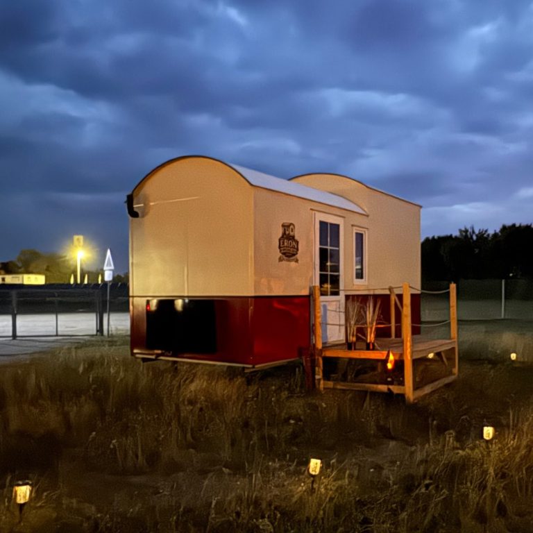 Tiny House mit rotem Streifen in der Dämmerung. Drumherum leuchten kleine Lampen im Gras.