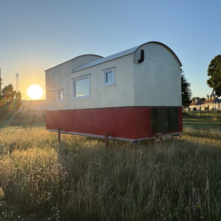 Tiny House mit rotem Streifen nach Sonnenaufgang auf einer Wiese.