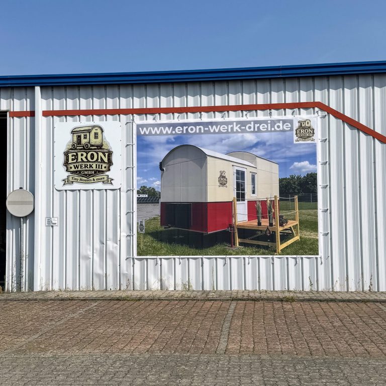 Ein großes Plakat mit einem Tiny House mit rotem Streifen und Runddach hängt an einer Hallenaußenwand. Daneben hängt ein Schild mit Logo.