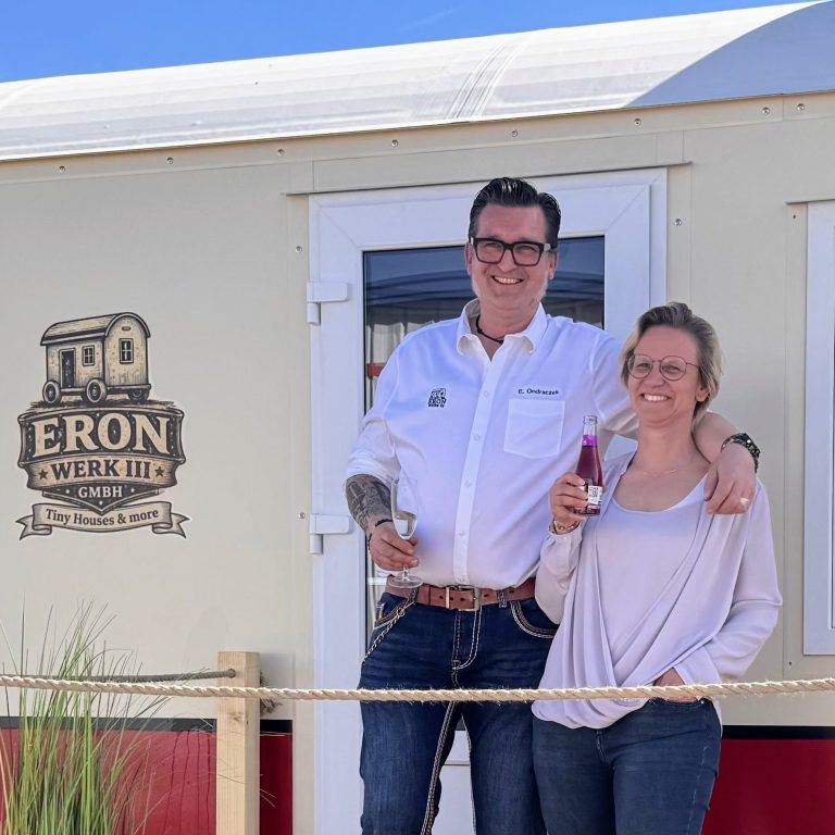 Erik Ondraczek & Eva Preetz Eine Frau und ein Mann stehen lächelnd vor einem Tiny House. Beide halten ein Getränk in der Hand.