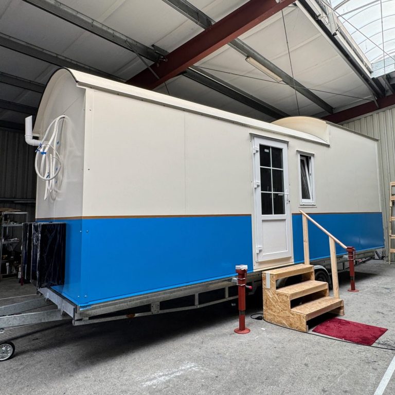 Tiny House mit blauem Streifen in der Produktionshalle. Treppe mit rotem Teppich davor.